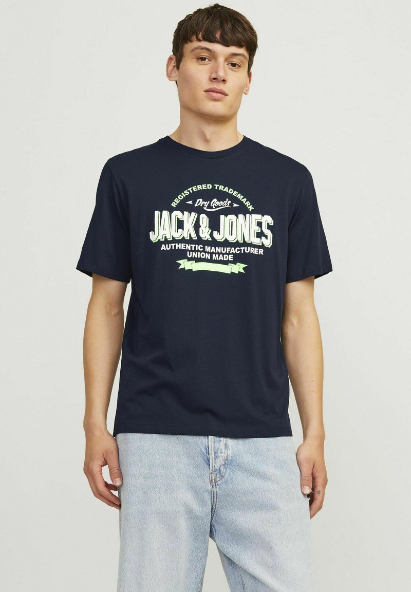 Tamsiai mėlynas medvilninis marškinėliai su dideliu baltu ir žaliu grafinio teksto dizainu, kuris ant priekio rašo "JACK & JONES". Trumpomis rankovėmis.