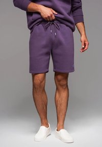 Fialové bavlněné šortky s elastickým pasem a šňůrkou, s bočními kapsami a nášivkou na lemu, nošené s bílými slip-on botami.