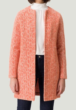 Manteau texturé à ouverture frontale en corail avec un design en boucle. Manches longues et coupe droite, comportant deux poches avant, porté sur un haut blanc.