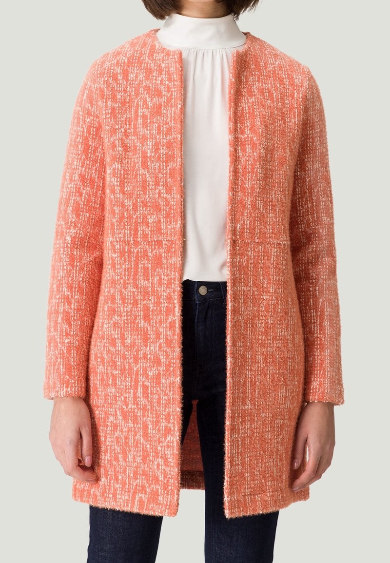 Textureret, åben-front frakke i koralfarve med et løkkedesign. Lange ærmer og lige snit, med to forlommer, båret over en hvid bluse.