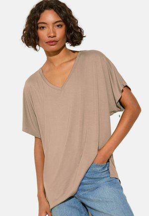 Frau mit kurzen, lockigen Haaren, die ein lockeres beiges V-Ausschnitt-T-Shirt mit kurzen Ärmeln und blaue Jeans trägt, eine Hand in der Tasche.