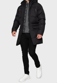 Giacca a vento nera con cappuccio, chiusura con zip, tasche laterali e finitura testurizzata. Indossata sopra un maglione grigio e pantaloni neri, abbinata a sneakers bianche.