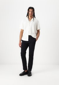 Camisa blanca de manga corta con botones y acabado texturizado, combinada con pantalones oscuros de corte slim y mocasines negros, de pie contra un fondo liso.