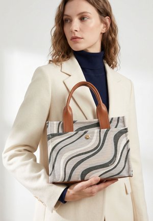 Donna che tiene una borsa beige con manici marroni e motivi astratti a onde nere e grigie, indossa un blazer crema e un dolcevita blu navy.