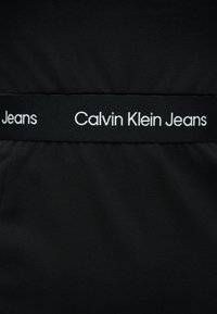 Μαύρη υφασμάτινη ζώνη μέσης με λευκό κείμενο "Calvin Klein Jeans" επαναλαμβανόμενο οριζόντια.