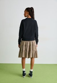 Felpa nera con maniche raglan abbinata a una gonna a balze a quadri. Sneakers alte bianche con accenti scuri, in piedi su un pavimento verde.