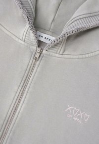 Lichtgrijze zip-up hoodie met een geribde zoom en capuchon, voorzien van een roze geborduurde tekst "XOXO OH APRIL" aan de linkerkant. Soepele stoffen textuur.