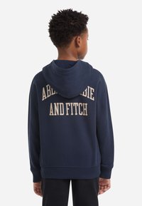 Ναυτικό μπλε φούτερ με μεγάλο μπεζ λογότυπο στην πλάτη. Διαθέτει κουκούλα με κορδόνι, ραμμένα μανίκια και τελείωμα. Μαλακή υφή, περιστασιακή εφαρμογή.