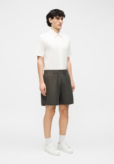 Νεαρός άνδρας με σγουρά μαλλιά, που φοράει λευκό κοντομάνικο πουκάμισο με γιακά, σκούρες γκρι βερμούδες με ελαστική μέση, λευκές κάλτσες και λευκά αθλητικά παπούτσια.