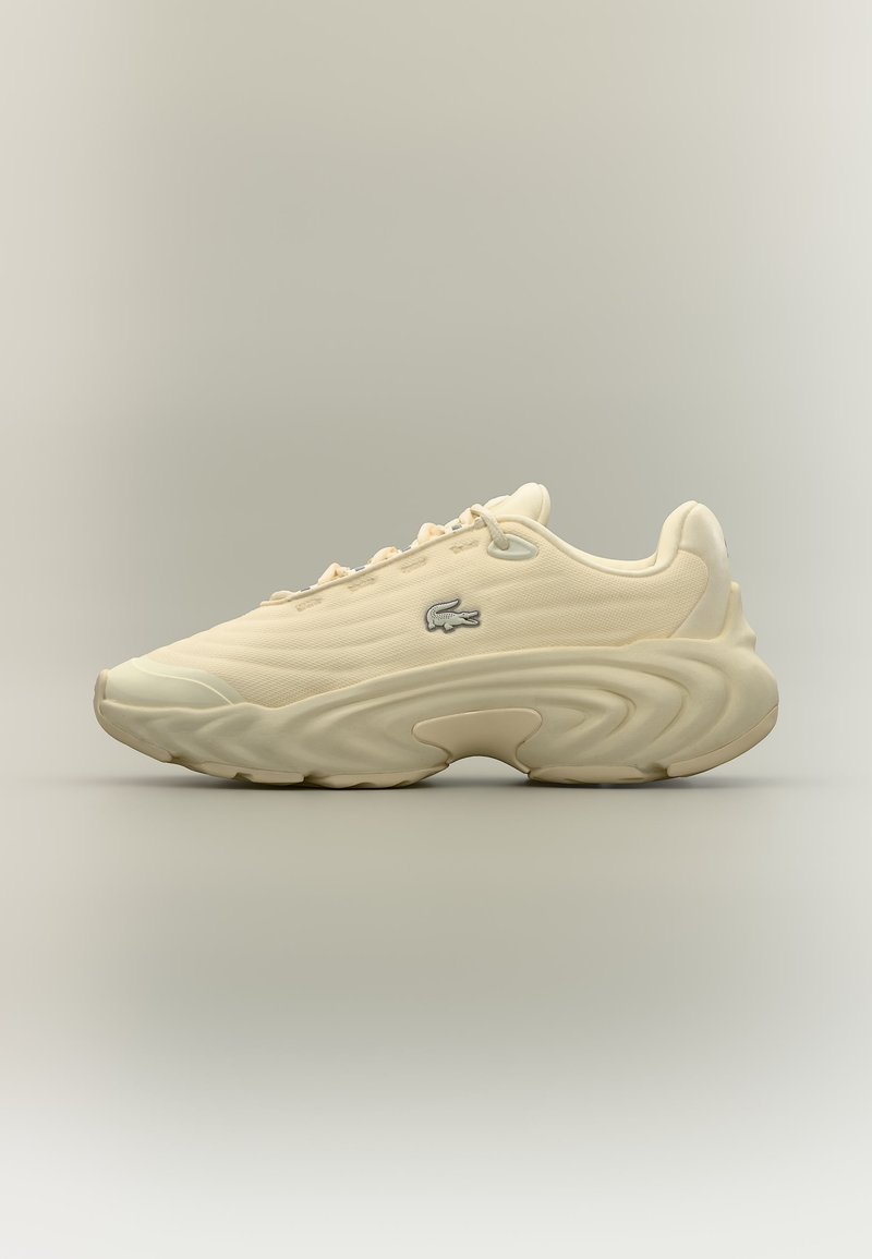 Sneaker beige chiaro con suola spessa ondulata, design con lacci e piccolo logo coccodrillo sul lato, esposta su sfondo neutro.