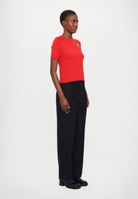 Rotes kurzärmeliges Strickoberteil mit rundem Ausschnitt, verziert mit einem dekorativen Patch. Kombiniert mit hochgeschnittenen schwarzen Hosen und Plateauschuhen.