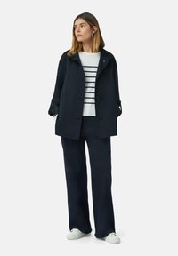 Marineblauwe jas met knopen en opgerolde mouwen, gecombineerd met een wit gestreept sweater en donkere, wijde broek, aangevuld met witte sneakers.