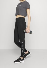 Zwarte high-waisted leggings met het "Calvin Klein" logo in het wit. Vergezeld van een donkergrijze cropped t-shirt en zwarte sportschoenen.