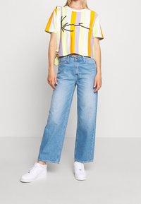 Lyhyt t-paita, jossa on pystysuorat raidoitukset oranssina, keltaisena, vihreänä ja purppurana, mustalla käsinkirjoitetulla logolla. Yhdistettynä väljälahkeisiin vaaleansinisiin farkkuihin ja valkoisiin lenkkareihin.