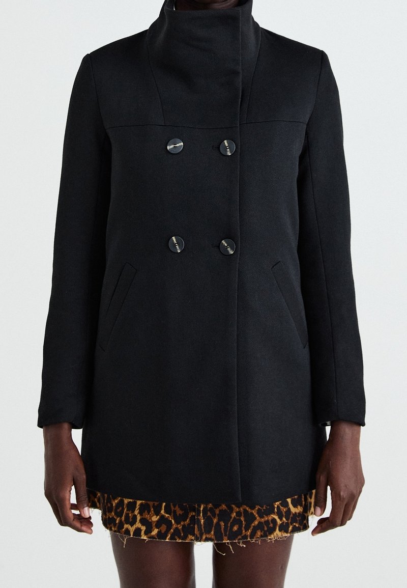Černý kabát z vlněné směsi s vysokým límcem, dvouřadým zapínáním, kulatými knoflíky a bočními kapsami. Hladká textura, přiléhavý silueta.
