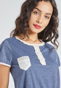 Una joven con cabello rizado lleva una camisa azul con un cuello de encaje y un bolsillo decorativo, sonriendo a la cámara.