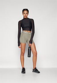 Crna dugih rukava majica, maslinasto zelene kratke hlače s pojasom "NIKE PRO", crne sportske tenisice, drži crnu bocu za vodu. Jednostavna pozadina.