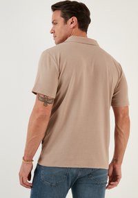 Beige kortærmet polo-shirt med en tekstureret stof. Har en klassisk krave og en lige snit, parret med blå jeans.
