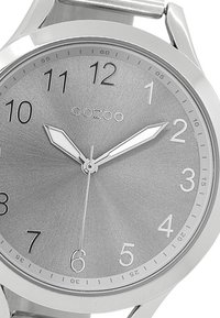 Montre-bracelet en argent avec un bracelet métallique, au design rond simple, grands chiffres et marqueurs d'heure minimalistes. Présente une texture brossée sur le cadran.