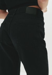 Jean noir en denim à taille haute, avec une patch au dos, deux poches arrière et une finition textile texturée.