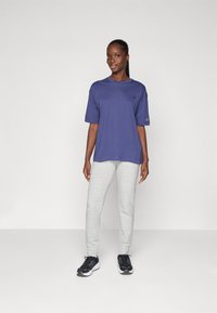 Maglietta di cotone viola con maniche corte e logo, abbinata a pantaloni della tuta grigi e sneackers nere con suole bianche.