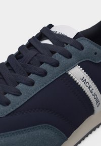 Σκούρο μπλε αθλητικό παπούτσι με γκρι σουέντ λεπτομέρειες, σκούρα μπλε κορδόνια, λευκή πλευρική ρίγα και λογότυπο "JACK&JONES" στην πλευρική επιφάνεια.