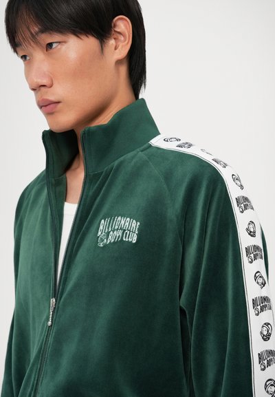 Casaco de veludo verde com fecho zíper, apresentando um colarinho alto, riscas laterais brancas com logotipo repetido e bordado do logotipo na frente. Textura suave.