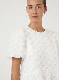 Λευκό μπλουζάκι φτιαγμένο από επίπεδα υφάσματα με σχέδιο ροζέτας από λουλούδια. Οι φουσκωτοί ώμοι και η στρογγυλή λαιμόκοψη ενισχύουν την υφή και την τρισδιάστατη εμφάνισή του.