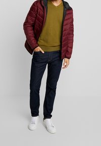 Muž v burgundské péřové bundě přes olivově zelený svetr, tmavých modrých džínách a bílých teniskách, stojící s jednou rukou v kapse.