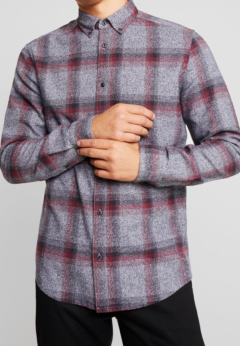 La camicia di flanella grigia presenta un motivo a quadri rosso e nero, colletto con bottoni e maniche lunghe. Il tessuto appare morbido e texturizzato.