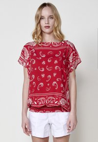 Top rosso a maniche corte con motivi bianchi in paisley e floreali, dal fit morbido e scollo a barca. Abbinato a pantaloni corti bianchi.