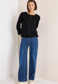 Schwarzes Langarmoberteil mit rundem Ausschnitt und Schlüssellochdetail, kombiniert mit weit geschnittenen blauen Jeans und cremefarbenen Sneakern.