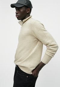 Maglione beige con zip e colletto a imbuto, con motivo testurizzato, polsini e orlo a coste. Indossato con jeans neri e un berretto di pelle nera.