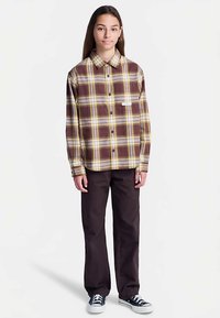 Hnedo-žltá košeľa s kockovaným vzorom, dlhými rukávmi, zapínaním na gombíky vpredu a náprsným vreckom, v spojení s tmavými nohavicami a teniskami.