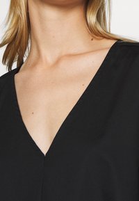 Černý top s hlubokým výstřihem do V, vyrobený z hladké látky. Má čisté linie a minimalistický design, bez viditelných vzorů nebo ozdob.