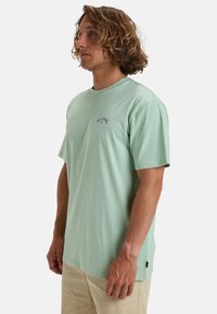 Lichtgroen T-shirt met korte mouwen en ronde halslijn, voorzien van een paarse logo op de borst. Zachte stof met een losse pasvorm, gecombineerd met een beige broek.