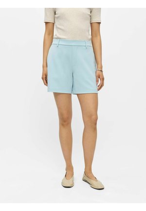 Vrouw die lichtblauwe op maat gemaakte shorts draagt, beige top met korte mouwen, beige platte schoenen en een zilveren armband aan haar rechterpols.