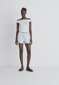 Weißes schulterfreies Top mit geraffter Optik, kombiniert mit hellen gestreiften Shorts. Das Model trägt flache schwarze Sandalen und steht vor einem schlichten Hintergrund.