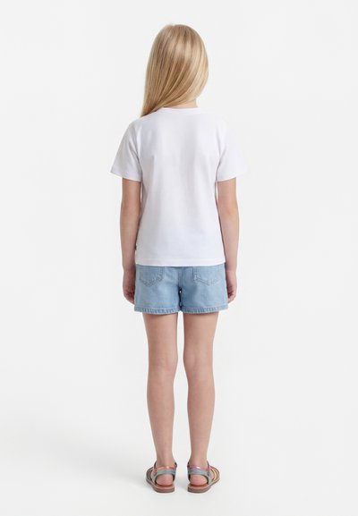 Παιδί με μακριά ξανθά μαλλιά στέκεται με την πλάτη γυρισμένη, φοράει απλό λευκό μπλουζάκι, ανοιχτό μπλε τζιν σορτς και πολύχρωμα σανδάλια.