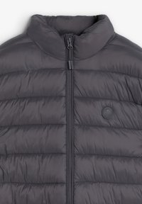 Grå puffervest lavet af nylon, med høj krave, frontlynlås, horisontalt quiltet mønster og et rundt logo patch på brystet.