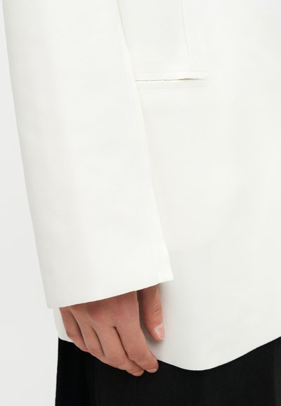 Κοντινό πλάνο ενός χεριού που ακουμπά στο πλάι, φορώντας ένα λευκό σακάκι με μακριά μανίκια και τσέπη με καπάκι, και μαύρο παντελόνι.