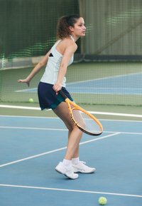 Tennisser in een lichtblauw tanktop en marineblauwe shorts, met een oranje racket, die zich voorbereidt om een tennisbal te slaan op een blauwe baan.