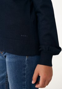 Marinblå stickad tröja med ribbade ärmslut och nederkant. Har subtil "MCXX"-logotyp på den nedre delen. Kombineras med blå denimjeans.