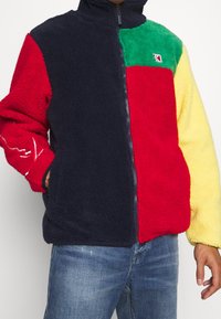 Fleecová bunda s barevným blokovým vzorem v námořnické modři, červené, zelené a žluté barvě. Celopropínací zip s vysokým límcem a bočními kapsami. Měkká textura.