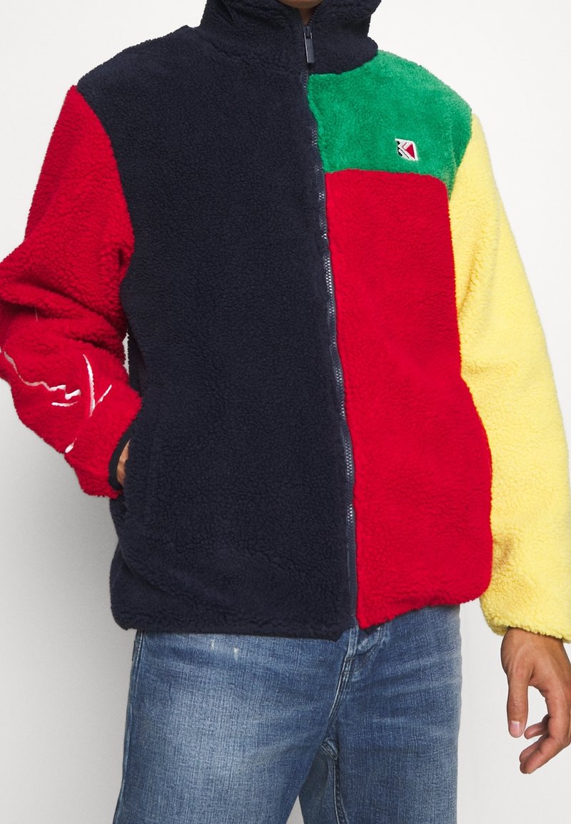 Fleecová bunda s barevným blokovým vzorem v námořnické modři, červené, zelené a žluté barvě. Celopropínací zip s vysokým límcem a bočními kapsami. Měkká textura.