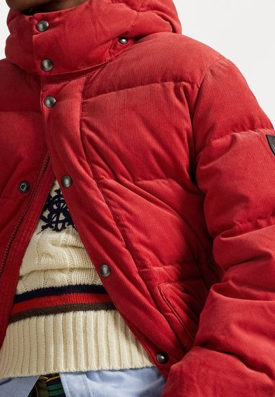 Jachetă roșie din țesătură corduroy, cu glugă, având nasturi tip snap și un model cu cusături. Sub ea este un pulover crem cu dungi orizontale.