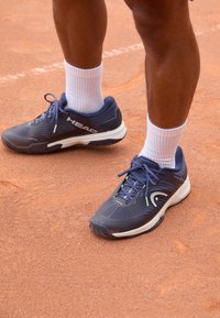 Kék sportcipők texturált felsőrésze és fehér talpa lime zöld kiegészítőkkel és fekete barázdával, fehér bordás zoknikkal viselve, agyagpályán.