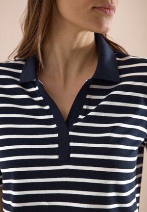 Mujer con camiseta polo de rayas horizontales azul marino y blanco con cuello abierto, mostrada desde el cuello hasta la parte superior del torso.