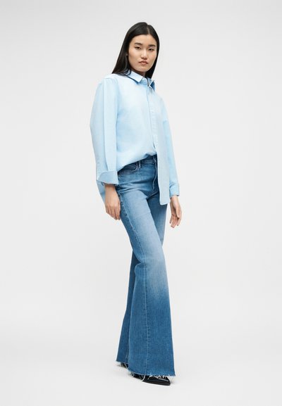 Žena stojaca, oblečená v voľnej svetlomodrej košeli na gombíky a v modrých vysoko pásových rozšírených džínsoch s čiernymi špicatými topánkami na bielom pozadí.