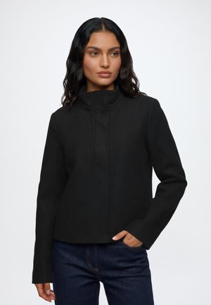 Kvinde med langt mørkt hår iført en sort højhalset jakke og mørkeblå jeans, med den ene hånd i lommen og kigger til siden.